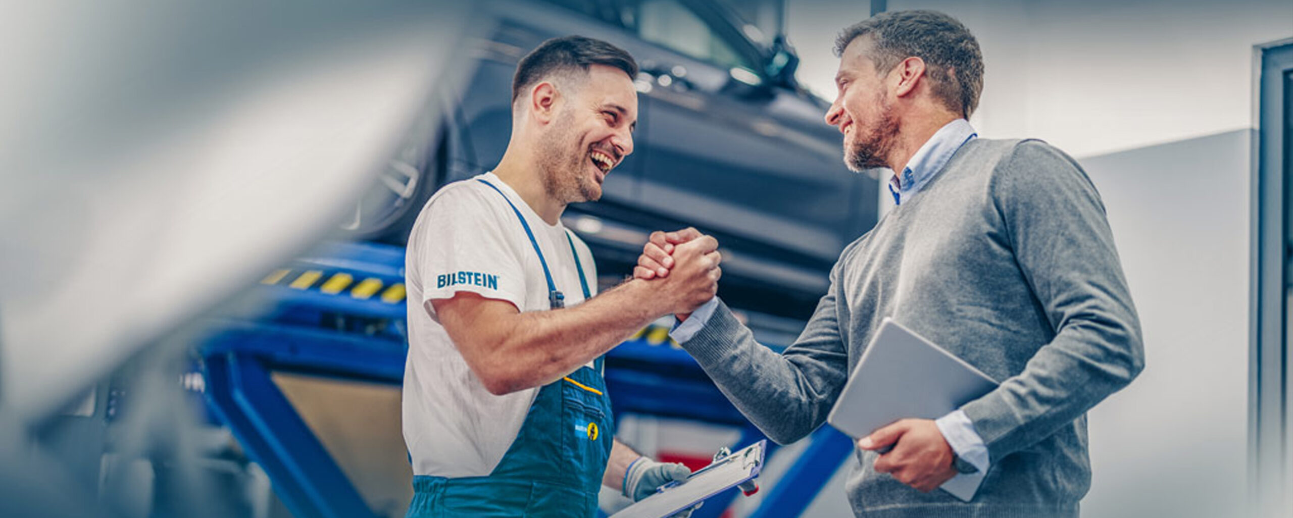 Handschlag zwischen Mechaniker und Mann im Hemd mit Werkstatt im Hintergrund